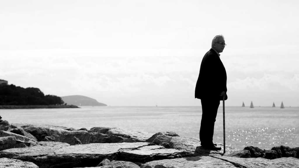 Elderly man with cane stands on rocky coastline, gazing at sea.
